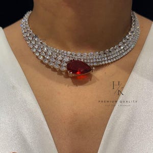 Op de afbeelding: Een sprankelende ketting met een grote, druppelvormige rode edelsteen hanger. De ketting is bezet met talrijke kleine, heldere edelstenen. De tekst "PREMIUM QUALITY Real Life Diamonds" is ook zichtbaar.