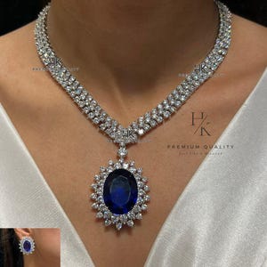 Op de afbeelding: Een luxe sieradenset bestaande uit een ketting, ring en oorbellen. De ketting heeft een grote, ovale, diepblauwe edelsteen hanger, omringd door kleinere, heldere stenen. De ring en oorbellen weerspiegelen dit ontwerp. De set is gemarkeerd met "Premium Quality."