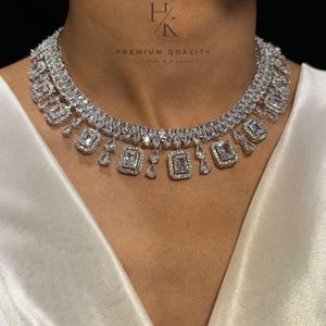 Peut inclure: Un collier argenté étincelant, orné de nombreux cristaux transparents. Le design présente des pierres carrées et en forme de goutte, disposées symétriquement. L'image affiche également le texte "PREMIUM QUALITY" et "Feel Like a Diamond".
