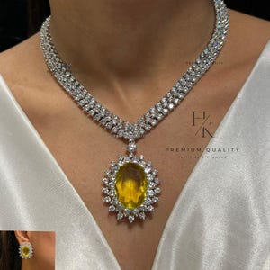 Op de afbeelding: Een luxe sieradenset bestaande uit een ketting, oorbellen en een ring. De ketting heeft een dubbelstrengs ontwerp met talrijke kleine, heldere stenen. Een grote, ovale gele stenen hanger is het middelpunt. De oorbellen en ring weerspiegelen het ontwerp van de hanger.