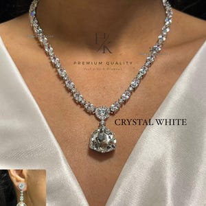 Pode incluir: Um colar de tom prateado com uma série de pedras claras de corte oval e um grande pingente triangular facetado no centro. O texto HK PREMIUM QUALITY Feel Like A Diamond e CRYSTAL WHITE N-1540 é visível na imagem.