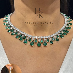 Op de afbeelding: Een zilverkleurige ketting en bijpassende oorbellenset met smaragdgroene, druppelvormige edelstenen en heldere, ronde accentstenen. De ketting heeft een trapsgewijs ontwerp en de oorbellen zijn een lineaire druppelstijl. De tekst "PREMIUM QUALITY Feel Like A Diamond" is inbegrepen.