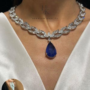 Pode incluir: Um colar prateado com um pingente central de pedra azul em forma de gota rodeado por pedras transparentes de corte baguette e marquise, com brincos a condizer. O texto HK PREMIUM QUALITY Feel Like A Diamond é visível.