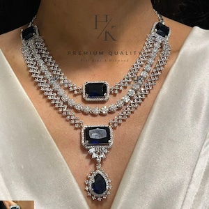 May include: A three-strand necklace and matching earrings set. The necklace features a series of clear, sparkling stones and large, dark blue rectangular center stones. The earrings have a similar design. The text "PREMIUM QUALITY" is visible.