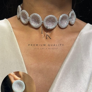 Op de afbeelding: Een sprankelende zilveren choker ketting en bijpassende oorstekers. De ketting heeft een reeks grote, ronde, met edelstenen bezette schakels. De oorbellen zijn een kleinere versie van de ketting schakels. De tekst "PREMIUM QUALITY" is zichtbaar.