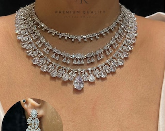 May include: A three-strand silver necklace with a large pear-shaped pendant. The necklace is adorned with numerous clear, sparkling stones. A matching earring is shown in the lower left corner. The text "PREMIUM QUALITY" is visible.