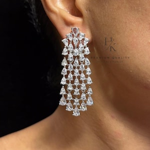 Könnte beinhalten: Ein langer, silberfarbener Kronleuchter-Ohrring mit einem kaskadierenden, mehrstufigen Design aus klaren, birnenförmigen Steinen. Der Ohrring ist in einen Metallrahmen gefasst und enthält den Text HK PREMIUM QUALITY im Hintergrund.