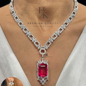 Pode incluir: Um colar de tom prateado com uma corrente de elos retangulares incrustados com pedras claras, centrado por um grande pingente de pedra vermelha retangular facetada rodeado por pedras claras menores. Texto HK PREMIUM QUALITY visível.
