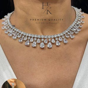 May include: A sparkling white necklace with teardrop pendants and matching earrings. The necklace features multiple rows of glittering stones, with the text "PREMIUM QUALITY Feel Like A Diamond" visible. Product code: N-1559-WHITE.