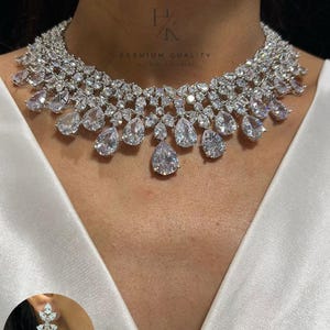 Peut inclure: Un collier argenté composé de plusieurs rangées de pierres transparentes à facettes, de forme ronde et en poire. Le design présente une goutte centrale en forme de poire. Une boucle d'oreille assortie est visible dans le coin inférieur.