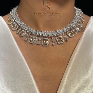 Op de afbeelding: Een sprankelende zilveren ketting versierd met heldere en amberkleurige edelstenen. De ketting heeft een gelaagd ontwerp met rechthoekige en druppelvormige stenen. De tekst "PREMIUM QUALITY" en "Feel like a Diamond" is zichtbaar.