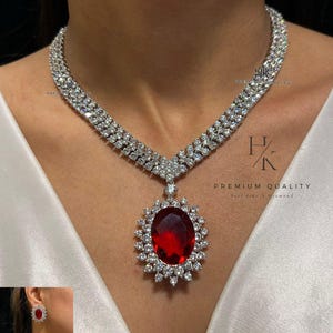 Op de afbeelding: Een oogverblindende ketting- en oorbellenset met een grote, ovale, dieprode edelsteen hanger, omringd door sprankelende, heldere stenen. De ketting heeft een dubbele rij ontwerp. De tekst "PREMIUM QUALITY" en "Feel like a diamond" is ook aanwezig.