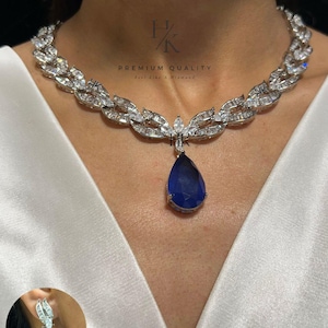 Op de afbeelding: Een zilverkleurige ketting met een bladvormig ontwerp, versierd met heldere kristallen en een grote, peervormige, diepblauwe edelsteenhanger. Bijpassende oorbellen met een vergelijkbaar ontwerp zijn ook zichtbaar. De tekst "PREMIUM QUALITY" staat op de achtergrond.