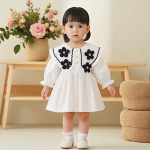 May include: A white dress with a large, scalloped collar adorned with black flower appliqués. The dress features long sleeves with ruffled cuffs and is worn by a young child. The background includes a wooden table and a vase of flowers.