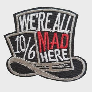 Pode incluir: Patch bordado preto e cinza em forma de chapéu alto. O texto "WE'RE ALL MAD HERE" está em branco, com "MAD" em vermelho. Os números "10/6" também são visíveis.