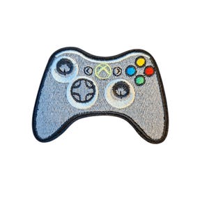 May include: An embroidered patch shaped like a video game controller. The controller is light gray with a black border. It features a central green logo, a directional pad, two analog sticks, and four colored action buttons in yellow, blue, red, and green.