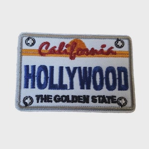 Könnte beinhalten: Gestickter Aufnäher in Form eines kalifornischen Nummernschilds. Der Aufnäher zeigt das Wort "California" in roter Schrift über "HOLLYWOOD" in blauen Blockbuchstaben. Der Text "THE GOLDEN STATE" steht darunter. Eine gelb-orangefarbene Sonnengrafik ist ebenfalls vorhanden.