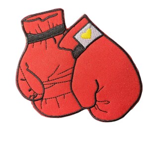 May include: Embroidered patch depicting two red boxing gloves with black trim. A small white square with a yellow heart is visible. The patch is designed for ironing or sewing onto fabric.