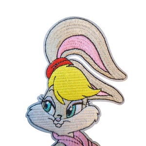 May include: Embroidered patch of a cartoon bunny with yellow hair, a red bow, and a pink scarf. The bunny has light blue eyes and a light tan face and ears. The patch is outlined in a light grey colour.