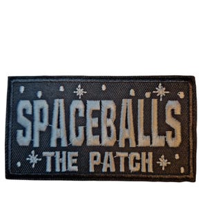 Peut inclure: Un écusson rectangulaire noir avec les mots "SPACEBALLS" brodés en gris clair. En dessous, le texte "THE PATCH" est également brodé en gris clair. Des éclats d'étoiles décoratifs sont brodés autour du texte.