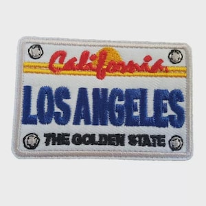 Può includere: Toppa ricamata a forma di targa californiana. La toppa presenta le parole "LOS ANGELES" in blu, "California" in rosso e "THE GOLDEN STATE" in nero. Un disegno di sole giallo è sopra il testo blu.