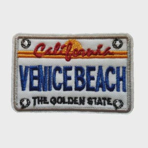 Pode incluir: Patch bordado em forma de placa de carro da Califórnia. O patch apresenta as palavras "Venice Beach" em azul, "California" em vermelho e "The Golden State" em preto. O fundo é branco com um desenho de sol amarelo e laranja.