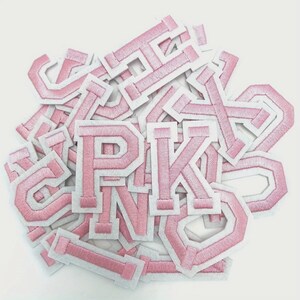 Op de afbeelding: Een verzameling roze geborduurde letterpatches met witte randen. De letters zijn in een klassieke collegiale stijl, met een getextureerde, gestikte uitstraling. De patches zijn verspreid, waardoor een visueel aantrekkelijke opstelling ontstaat.