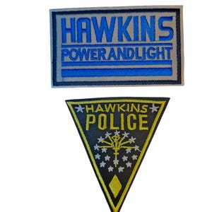 May include: Two embroidered patches. The top patch is rectangular with the text "HAWKINS POWER AND LIGHT" in blue. The bottom patch is a black triangle with "HAWKINS POLICE" and a gold emblem.