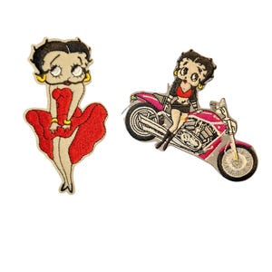 Peut inclure: Deux écussons brodés représentant Betty Boop. L'un montre Betty dans une robe rouge, l'autre sur une moto rose. Les deux écussons ont un fond crème et des contours noirs.