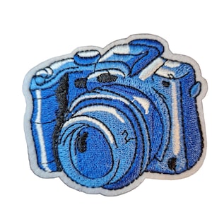 May include: Embroidered patch of a blue camera with white highlights. The camera lens is detailed with a circular design and the number "2". The patch has a white border.
