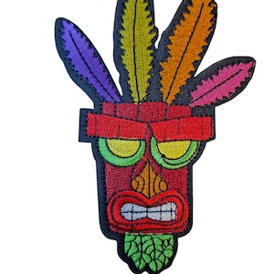 May include: Embroidered patch featuring a stylized tribal mask with a red face, green eyes, and a wide, toothy grin. The mask is adorned with colorful feathers in purple, yellow, orange, and pink. The patch has a black border.