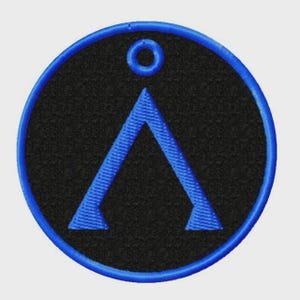 Puede incluir: Un parche bordado circular con fondo negro y borde azul brillante. Centrado en el parche hay un símbolo triangular azul en forma de cheurón que apunta hacia arriba, situado directamente debajo de un pequeño icono de anillo circular azul.
