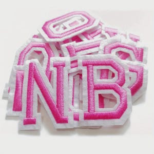 Op de afbeelding: Een stapel witte stoffen patches geborduurd met roze letters. De letters zijn omrand met roze draad en hebben een witte rand. De patches zijn in verschillende vormen en maten, met sommige letters gedeeltelijk zichtbaar.