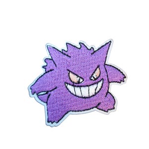 Può includere: Una toppa ricamata viola raffigurante un fantasma dei cartoni animati con un ampio sorriso e denti affilati. Il fantasma ha tratti appuntiti e occhi arancioni, delineati in nero. La toppa è bordata da un contorno bianco.