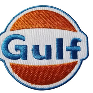Könnte beinhalten: Gestickter Aufnäher mit dem Gulf-Logo. Das Design umfasst einen weißen Hintergrund, einen blauen Rand, das Wort "Gulf" in Blau und orangefarbene Halbkreise. Der Aufnäher ist wahrscheinlich für Kleidung oder Accessoires bestimmt.