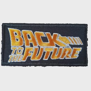 Op de afbeelding: Geborduurde patch met de tekst "BACK TO THE FUTURE" in goud en wit op een zwarte rechthoekige achtergrond. De patch heeft een retro-ontwerp, met de woorden gestapeld en gestileerd.