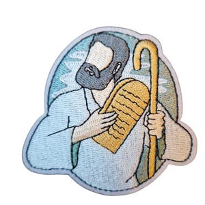 May include: Embroidered patch featuring a cartoon depiction of a bearded figure holding a tablet and staff. The figure is dressed in light blue robes, with a gray beard and hair. The tablet is yellow, and the staff is gold. The patch has a white border and a blue background.