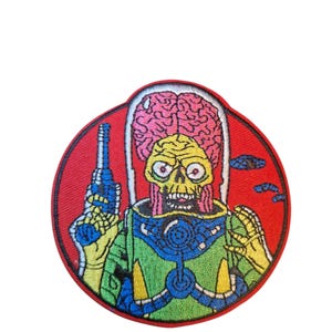Op de afbeelding: Geborduurde patch met een cartoon alien met een groot brein, een geel schedelachtig gezicht en een groen en blauw uniform. De alien houdt een blauw straalgeweer vast tegen een rode cirkelvormige achtergrond.