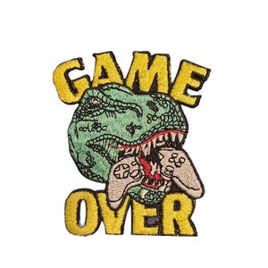 Op de afbeelding: Geborduurde patch met een groene dinosauruskop die een videogamecontroller bijt. De woorden "GAME OVER" staan in het geel, boven en onder de dinosaurus.