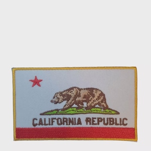 May include: Embroidered patch depicting the California Republic flag. The patch features a light blue background with a brown bear, green grass, and a red star. The words "CALIFORNIA REPUBLIC" are embroidered in dark letters.