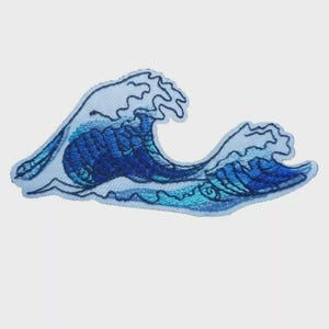 May include: Embroidered patch depicting a stylized ocean wave in various shades of blue. The wave design has a white outline and detailed stitching, giving it a textured look. Ideal for customising clothing or accessories.