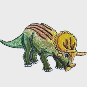 May include: Embroidered patch of a triceratops dinosaur. The patch features a green body with yellow and orange stripes on its back. The dinosaur has a yellow frill and horns, with white accents on its feet.