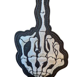 Könnte beinhalten: Gestickter Aufnäher mit einer weißen Skeletthand, die den Mittelfinger zeigt. Der Aufnäher hat einen schwarzen Rand und detaillierte Knochenstrukturen. Ein einzigartiges Accessoire für Jacken, Taschen oder Kleidung.