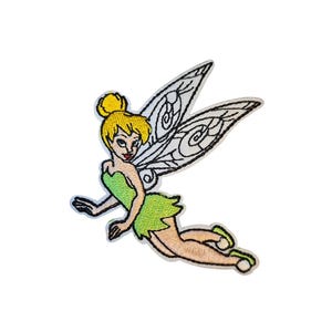 May include: An embroidered patch featuring a fairy character with blonde hair in a bun, wearing a green dress and shoes. The fairy has large, detailed white wings with black outlines and is depicted in a flying pose with a light skin tone.