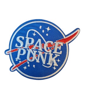 May include: Circular embroidered patch with a blue background and the words "SPACE PUNK" in white. The patch features a red swoosh design and white stars, reminiscent of a NASA logo. Ideal for jackets or bags.