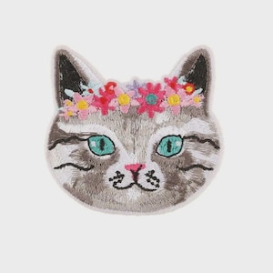 May include: Embroidered patch of a cat's face wearing a floral crown. The cat has grey and white fur, turquoise eyes, and a pink nose. The flowers in the crown are pink, yellow, and blue. The patch is set against a white background.