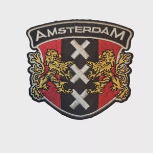 May include: Embroidered patch of the Amsterdam coat of arms. The shield-shaped patch features a red background, black and white stripes, and two gold lions. The word "AMSTERDAM" is arched across the top in white lettering.