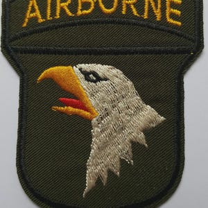 Może przedstawiać: Haftowana naszywka z napisem "AIRBORNE" złotą nicią nad głową orła. Orzeł ma żółty dziób i czerwone usta, ze szczegółowymi białymi i szarymi piórami. Naszywka jest oliwkowozielona z czarną obwódką.