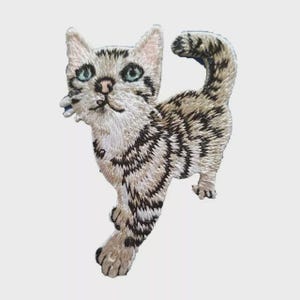 Peut inclure: Écusson brodé d'un chat marchant avec un motif rayé gris clair et noir. Le chat a les yeux bleus, un nez rose et un visage blanc. L'écusson est sur fond blanc.