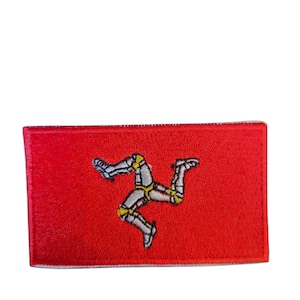 May include: A red rectangular patch embroidered with the Isle of Man emblem. The emblem shows three armored legs, joined at the thigh, with yellow detailing. The patch has a white border and is set against a red background.